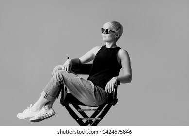 Monochrome Portrait Of Stylish Blonde Young Woman In Black Sun Glasses, Black Shirt And Jeans Sitting On Director Chair On  Wall Background 