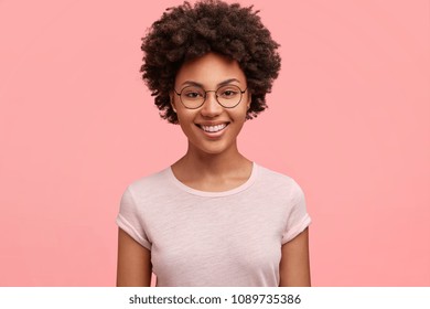 Monochrome shot of happy African American businesswoman with positive smile, has crisp dark hair, rejoices having weekend and good rest after hard working exhausting week, isolated on pink wall