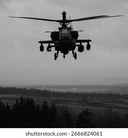 Monochrome, head‑on view of an Apache attack helicopter hovering over a training area with spinning rotors beneath a moody, overcast sky.