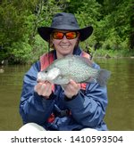 A monster black crappie fish being held horizontally by a smiling women angler in a hat and sunglasses on a brown water bay in spring time on a sunny day