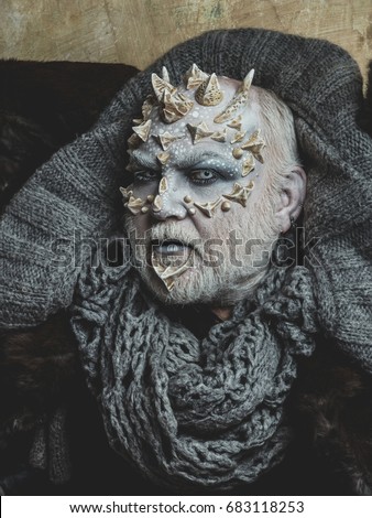 Monster face with white eyes, sharp thorns and warts. Demon head with grey collar on abstract beige wall. Horror and fantasy concept. Alien or sorcerer makeup. Man with dragon skin and beard.