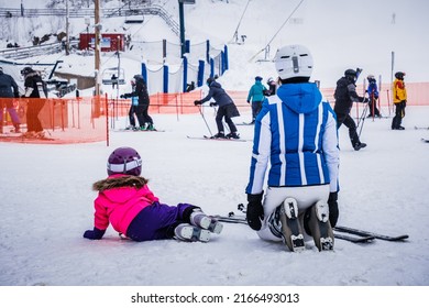 Mont Tremblant, Qc, Canada - January 18th 2021: Woman And Her Daughter, Wearing Ski Suits And Helmets, Sitting On The Snow Watching Skiers At The Bottom Of The Tremblant Ski Resort Slopes