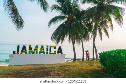 Montego Bay, Jamaica - July 2018: Couple kissing on beach, past a large sign of 'Jamaica' letters in the Jamaican flag, with beach and palm tree background.