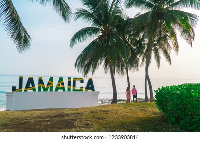 Montego Bay/Jamaica - July 2018: Couple walking on beach, past a large sign of 'Jamaica' letters in the Jamaican flag, with beach and palm tree background.