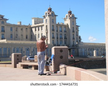 MONTEVIDEO, URUGUAY, March 22, 2016: Man Fishing In Parlasur, On The Waterfront Of The South Rambla In The Sun. In The Background The Mercosur Parliament Building In Front Of Ramirez Beach.