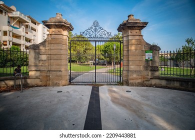 Montpellier, France - September 14th 2021 : Large Black Metal Door Opening Onto A Green Park With Lawns And Trees, A Person Sitting In The Grass On The Left, Tar In Front Of The Entrance, Blue Sky