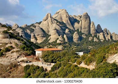 Montserrat Spain. The Mountains Behind The Benedictine Monastery. Small Building In Foreground With Groups Of People. Trees And Shrubs In Leaf. Blue Sky Some Cloud.