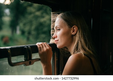 Mood Atmospheric Lifestyle Portrait Of Young Beautiful Blonde Hair Girl Looking Out Of Window From Riding Train. Pretty Teen Enjoying Beauty Of Nature From Moving Train Car In Summer. Travel Concept.