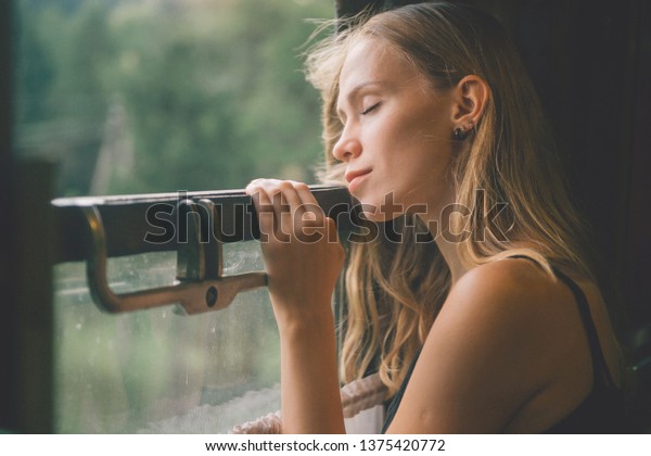 Mood atmospheric lifestyle portrait of young
beautiful blonde hair girl looking out of window from riding train.
Pretty teen enjoying beauty of nature from moving train car in
summer. Travel concept.