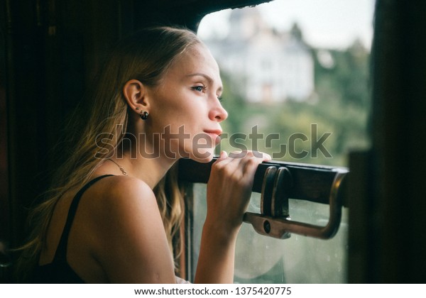 Mood atmospheric lifestyle portrait of young
beautiful blonde hair girl looking out of window from riding train.
Pretty teen enjoying beauty of nature from moving train car in
summer. Travel concept.
