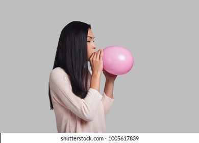 In a mood for a big holiday. Studio shot of attractive young woman in casual wear blowing up a pink balloon while standing against grey background