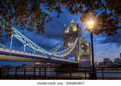 Moody Autumn View To The Tower Bridge Of London, UK, At Evening With Streetlight And Golden Leafs At The Trees