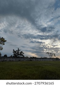 Moody, dramatic cloudy sky over a grassy field and a tall concrete wall at sunset or twilight. A touch of blue and yellow peeks through the dark, textured clouds.