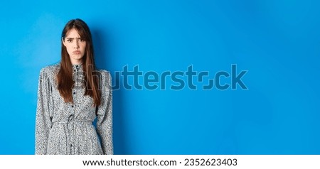 Moody frowning girl in dress looking at unfair thing, standing offended or upset against blue background.