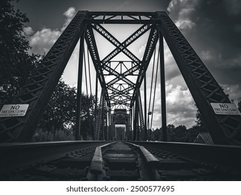 A moody, low-angle view of a steel truss railway bridge in Augusta, Georgia, with “No Trespassing” signs and dramatic clouds, emphasizing the structure’s imposing design in black and white. - Powered by Shutterstock