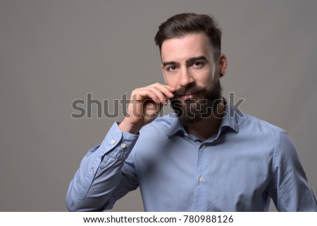 Moody portrait of young stylish bearded man twirling mustache and looking at camera over gray studio background with copyspace.