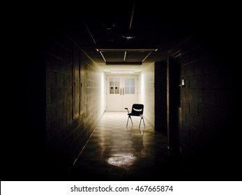 Moody, Shadowy Photo Of An Isolated School Chair - Placed At The End Of A Dark, Empty Hallway With A Single Source Of Light In The Background