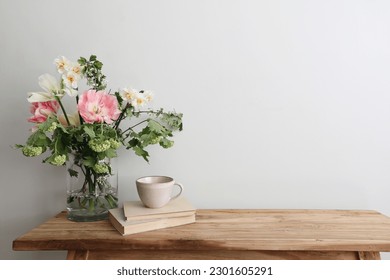Moody spring still life. Wooden bench, table composition with cup of coffee, tea and old books. Beautiful floral bouquet with white, pink tulips, daffodils. Hawthorn, green guelder rose flowers. Wall.