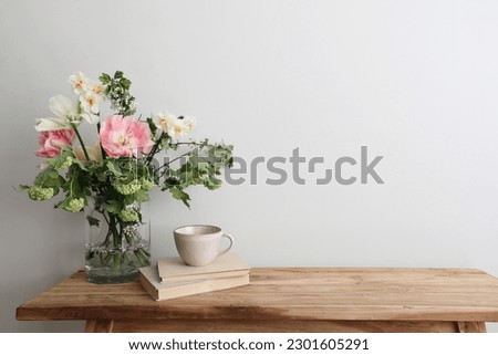 Moody spring still life. Wooden bench, table composition with cup of coffee, tea and old books. Beautiful floral bouquet with white, pink tulips, daffodils. Hawthorn, green guelder rose flowers. Wall.