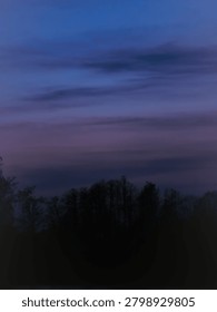 Moody twilight landscape featuring a dramatic sky with layered clouds transitioning through deep blues, purples, and soft magentas. The ethereal colors contrast against the dark, silhouetted treeline, capturing the serene atmosphere of dusk or dawn over a natural forest.