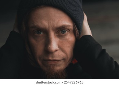 In a moody urban atmosphere, a young man with long hair in a beanie covers his ears, emulating a moment of introspection and vulnerability amidst the chaos. - Powered by Shutterstock