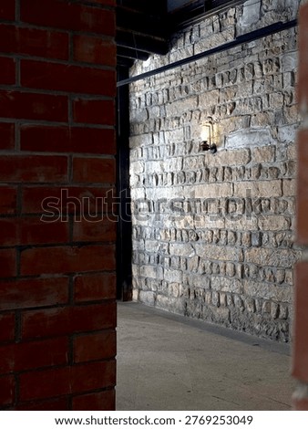 A moody, vertical composition showcasing the sharp corner where a dark red brick pillar meets a textured, dimly lit stone wall. The high-contrast lighting creates a mysterious and dramatic atmosphere, highlighting the raw, industrial beauty of historical building materials. This shot is perfect for themes related to mystery, historical cellars, or underground architectural tours. The minimal color palette of deep reds and stony greys provides a professional and sophisticated look for editorial pieces on urban exploration or masonry craftsmanship.