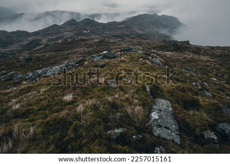 Moody view of Tarn Crag in the Lake District National Park in England on a cold day with dramatic clouds. Rocks and moss are in the foreground. 