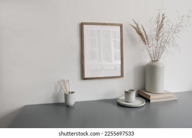 Moody wokspace still life. Pencils in ceramic holder. Vase with dry grass and cup of coffee. Blank vertical picture  frame mockup hanging on white wall background. Grey table background. Home office. - Shutterstock ID 2255697533