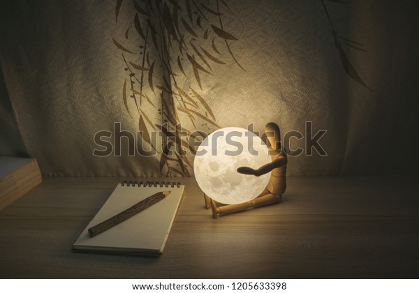 Moon lamp on desk, wooden doll hug\
the Moon. reading book on wooden table, with Chinese blinds,\
notebook, pencil and book, hipster style with copy space\
