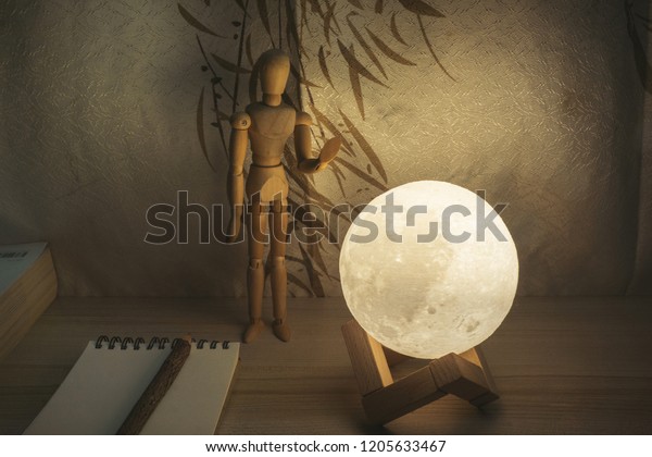 Moon lamp on desk with wooden doll hug the Moon.\
reading book on wooden table, with Chinese blinds, notebook, pencil\
and book.