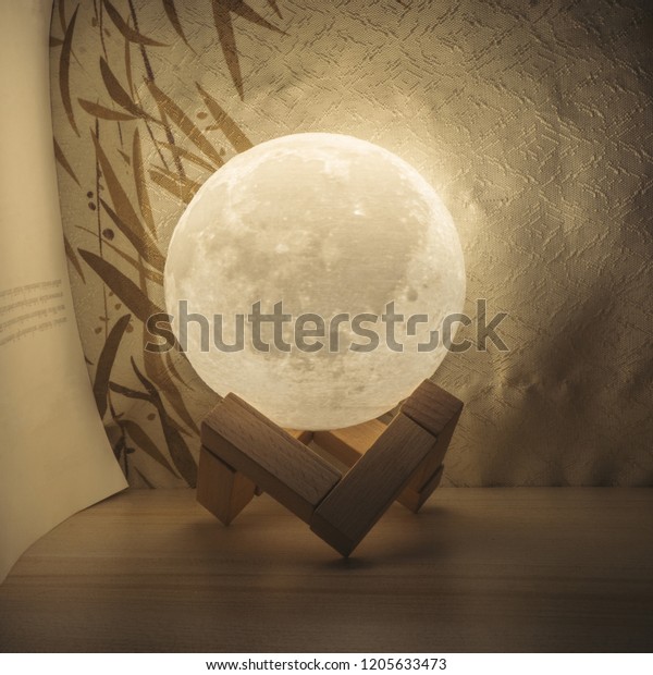 Moon lamp on\
desktop table, at night with warm light with book and blinds\
behind. moonlight on wooden table\
\

