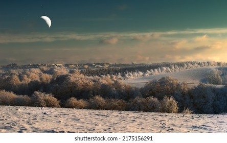 The moon in winter landscape. A half moon over snow covered bushes and shrubs in a field with tiny houses in the background. The Moon shining through clear clouds in dim sky - Powered by Shutterstock