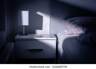 Moonlight in bedroom at night from window. Bed and table in dark room at dusk. Interior design in modern white apartment. Blue light from moon. Lamp on nightstand in empty home or in hotel or motel.