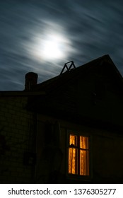 Moonlit Night With Floating Clouds Above A Village House With Light In The Window.