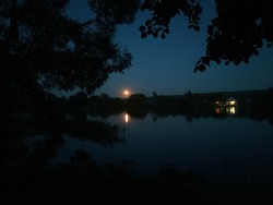 Moonrise Over The River On A Summer Windless Night. Trees Looming On The River, On The Other Side The Light From The Village Houses