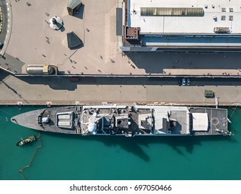 Moored Battleship In The Dock. On The Pier There Is A Building, People And Cars. Top View Photo. Horizontal.