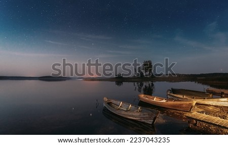 Moored Old Shabby Wooden Fishing Rowboats , Left Afloat On The Motionless Clear River Lake Water Next To Waterside. Amazing Glowing Stars Effects Above Lake. Night Starry Sky Soft Colors.