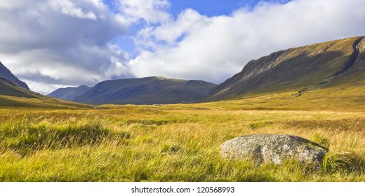 Moorland And Mountains In The Scottish Highlands Near Glencoe, Scotland, With Wild Flowers, Purple Heather And A Granite Boulder In The Foreground.