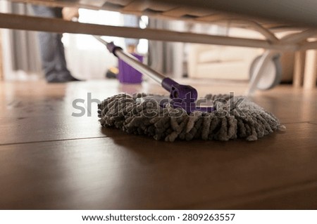 Mopping under sofa illustrates housework, hygiene and everyday home maintenance in a living room interior, linking the subject, setting and commercial concept in one factual moment.