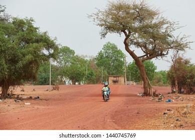 Mopti / Mali - May 19, 2018: African women riding motorcycle on dirt earth unpaved sandy road surrounded by trees and plastic garbage.