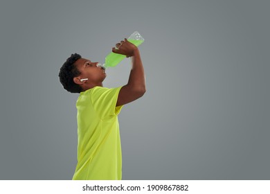 More Energy. Side View Of A Teenage African Boy Wearing Sports Clothes Drinking Energy Drink While Standing Against Grey Background, Web Banner With Copy Space For Text. Young People And Sport