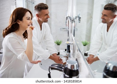 Morning Activities. Waist Up Of A Positive Couple In The Bathroom Where A Smiling Man Washing His Hands And Happy Woman Brushing Her Teeth