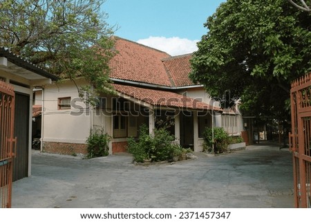 morning atmosphere on the veranda of an old house with yellowish wall paint and reddish orange roof tiles, there is a large red gate in front of the house and decorated with green trees 