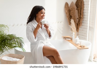 Morning Beauty Ritual. Happy African American Lady Drinking Coffee Sitting On Bathtub In Modern Bathroom Indoors. Wellness And Spa, Self Care And Pampering Concept. Side View