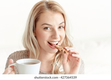 morning, breakfast, food and people concept - happy young woman with cup of tea eating cookie at home