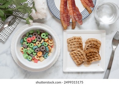 Morning breakfast spread with colorful cereal, mini waffles with syrup, and bacon strips served on white plates and bowls on a white marble backdrop, a spoon, fork, table napkin, florals, and a glass  - Powered by Shutterstock