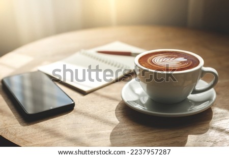 Morning in cafe, latte coffee in white cup witn smart phone and note book on wooden table with sunlight shines through, Close-up view