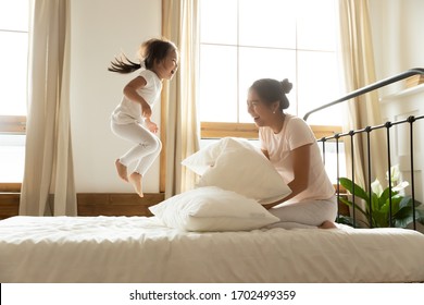 In morning carefree small daughter jump on bed while vietnamese mother laughing feels happy, asian ethnicity family in comfortable pyjamas wake up start new day positive mood enjoy active life concept