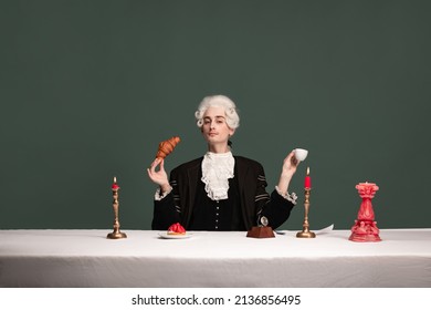 Morning coffee. Portrait of young elegant man in peruke and vintage jacket sitting at table isolated on dark green background. Retro style, comparison of eras concept.