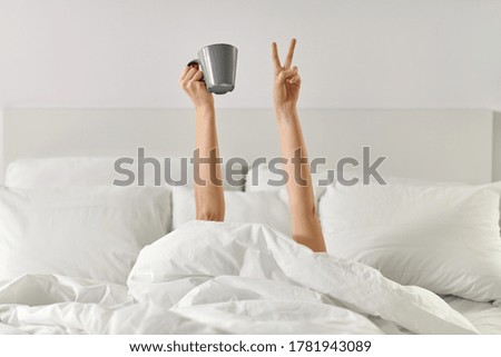 morning, comfort and people concept - young woman with cup of coffee lying in bed and showing peace hand sign at home bedroom
